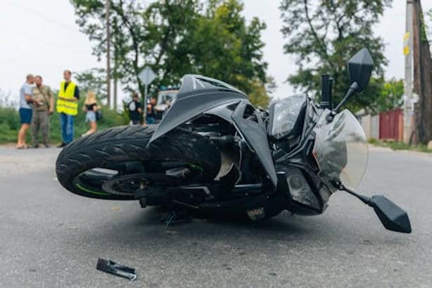 A black motorcycle lies on its side on a road after an accident, with debris scattered nearby and people standing in the background.