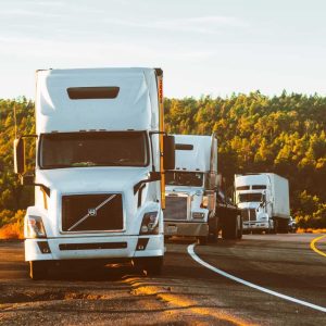 Three white semi-trucks parked along a roadside, with a forested hill in the background under a clear sky.