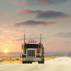 A red semi-truck drives on a deserted highway during sunset. The road stretches into the distance, lined with dry grass and a wire fence under a partly cloudy sky.