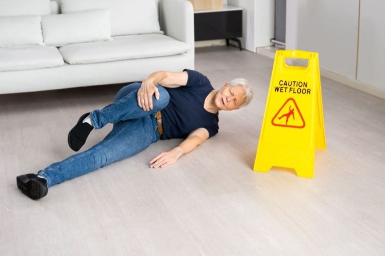 An older adult lies on the floor next to a yellow "Caution Wet Floor" sign in a living room setting.