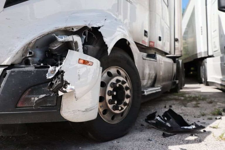 A close-up of a white semi-truck with visible front-end damage, including a broken bumper and detached parts on the ground.