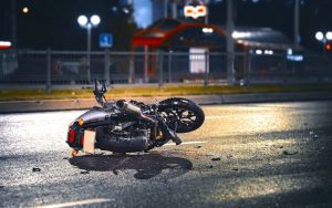 A motorcycle lies on its side on a wet street at night with scattered debris around; a blurred background shows streetlights and a bus stop.