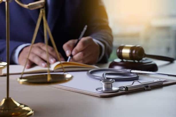 A person writes at a desk with a clipboard, stethoscope, gavel, and scales of justice, suggesting a legal and medical context.