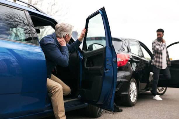 Two men exit their vehicles after a car accident involving a blue car and a black car on a city street.