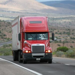 A red semi-truck drives on a highway through a desert landscape, with mountains visible in the distance.