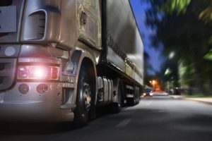 Close-up of a moving semi-truck on a road at dusk, with headlights on and blurred trees in the background.