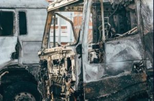 Two severely burned and charred vehicles, stripped of most external features, parked in an outdoor area with a building visible in the background.