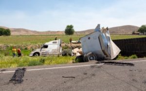 A white semi-truck has crashed off the road into a field, with its trailer damaged. Three people in safety vests stand nearby. Debris and tire marks are visible on the road.