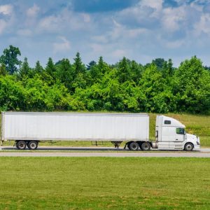 A white semi-truck with a long trailer drives on a road, surrounded by green grass and trees, under a partly cloudy sky.