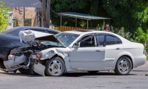 Two damaged cars on a street corner after a collision. The white car has a crumpled front, while the dark car is partially visible. Trees and buildings are in the background.