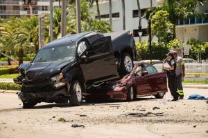 A black pickup truck is atop a red sedan after a collision. Two people in uniforms and a civilian are standing nearby. Various debris is scattered on the road.