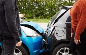 Two people stand near a collision between a blue car and a black car on a road with trees in the background.