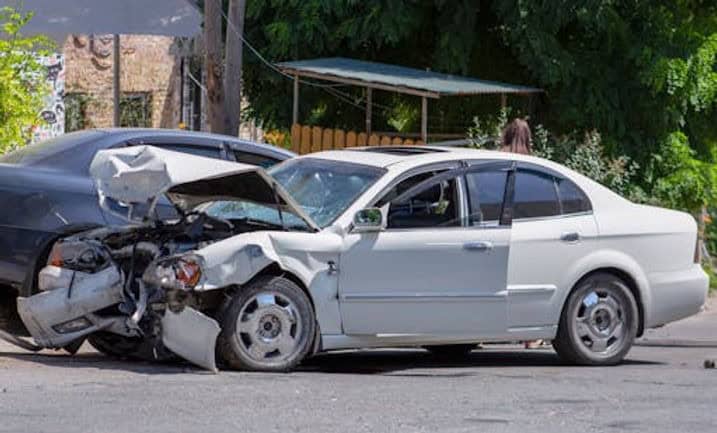 A white sedan with a severely damaged front end is involved in a collision with a black car on a street; debris is visible on the ground.