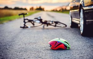 A bicycle and a helmet lie on the road near a parked car, suggesting a possible accident scene at sunset.
