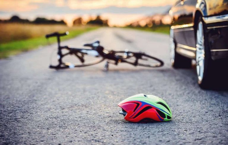 A bicycle and a helmet lie on the road near a parked car, suggesting a possible accident scene at sunset.