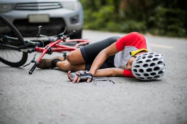 A cyclist wearing a helmet and gloves is lying on the road next to a fallen bicycle, with a car parked nearby.