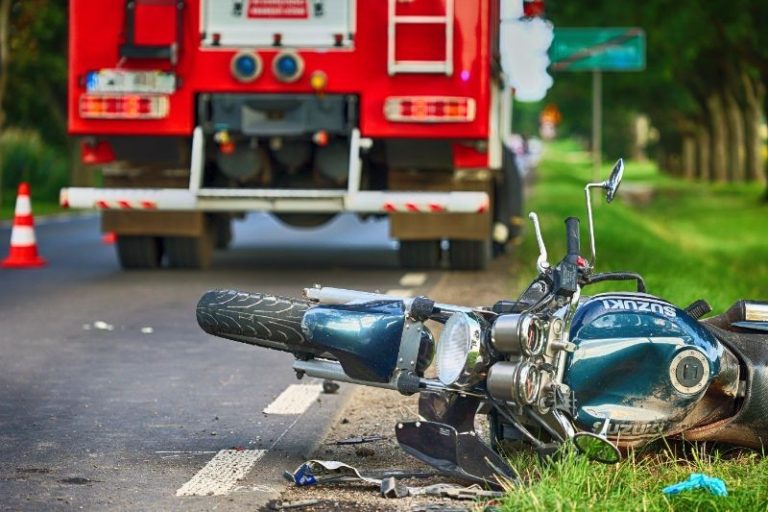 A motorcycle lies on its side on the road after an accident, with a fire truck and traffic cones positioned nearby.
