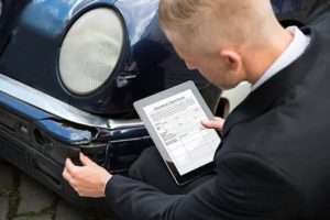A man in a suit examines a damaged car bumper while holding a tablet displaying an insurance claim form.