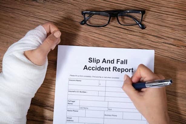 A person with a bandaged hand fills out a Slip and Fall Accident Report form on a wooden table, with glasses placed above the paper.