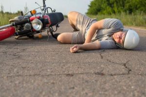 Person lying on the road in protective gear beside a fallen motorcycle; green shrubs in the background.