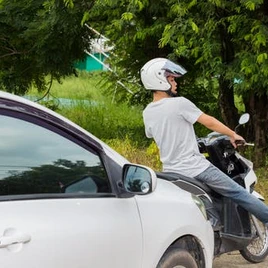 A person wearing a white helmet rides a black scooter near a parked white car, with green trees and grass in the background.