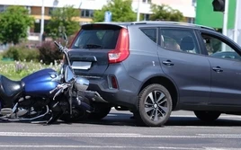 A blue motorcycle lies on its side after colliding with the rear of a gray SUV on a city street during daylight.