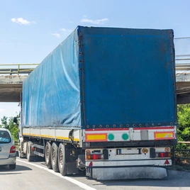 A large truck with a blue tarp is stuck under a low bridge, partially blocking the road while a car is parked nearby.