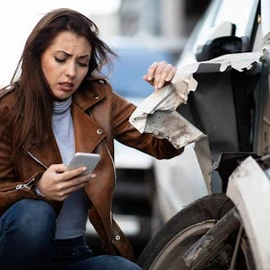 A woman kneels next to a damaged car, looking at her phone with concern. The car's front bumper is visibly dented and broken.