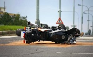 A motorcycle lies on its side in the middle of a road, with sand scattered around it and a caution sign visible in the background.