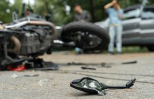 A damaged motorcycle and broken side mirror lie on the road, with a car and two people standing in the background.