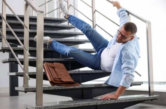A man wearing glasses falls backward on a staircase, one hand holding the railing and the other on a step, with a brown briefcase lying nearby.