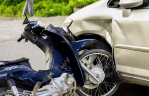 A damaged motorcycle is collided with the front side of a white car on a road.