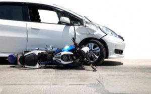 A damaged white car with a dented front side is next to a toppled blue and white motorcycle on a paved road.