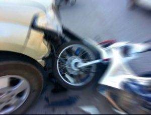 A blurred image of a crash between a beige car and a white motorcycle, showing the impact on the front wheel of both vehicles.