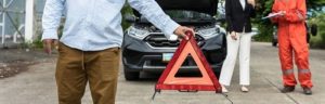 A person sets up a warning triangle in front of a car with an open hood, while two others converse by the vehicle.