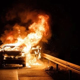 A car engulfed in flames on the side of a road at night, with fire coming from the front and interior, near a metal guardrail.