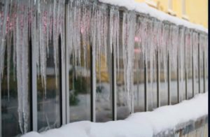 Long icicles hang from a snow-covered metal railing, with building windows and a faint reflection visible in the background.