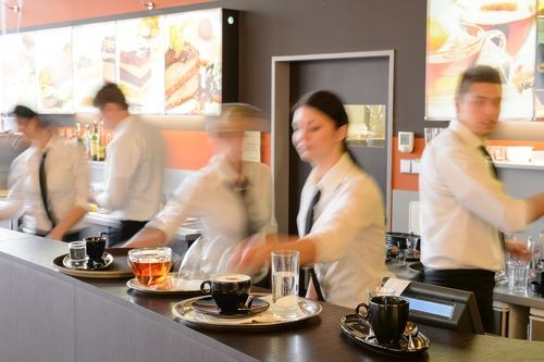 Waitstaff in white shirts and black ties work behind a counter, serving drinks including tea, coffee, and water in a busy restaurant or café setting.
