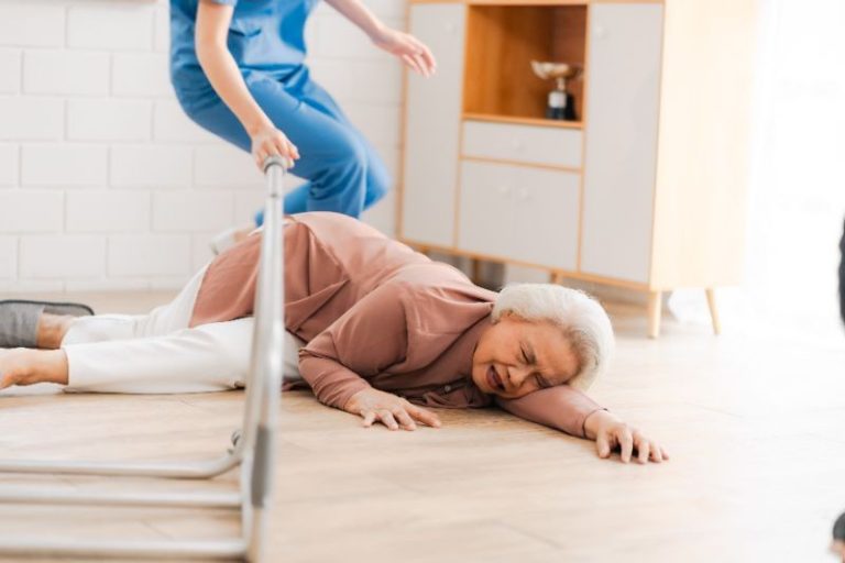 An older woman lying on the floor in pain next to a fallen chair, while a caregiver in scrubs approaches to help.