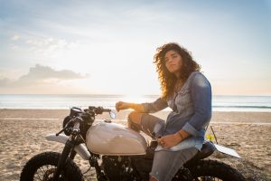 A woman in a denim shirt and jeans sits on a white motorcycle parked on a sandy beach at sunset, looking into the distance.