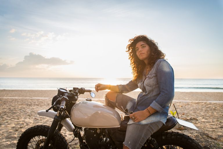 A woman in a denim shirt and jeans sits on a white motorcycle parked on a sandy beach at sunset, looking into the distance.
