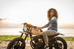 A woman with curly hair sits on a motorcycle parked on a sandy beach at sunset, looking toward the camera.
