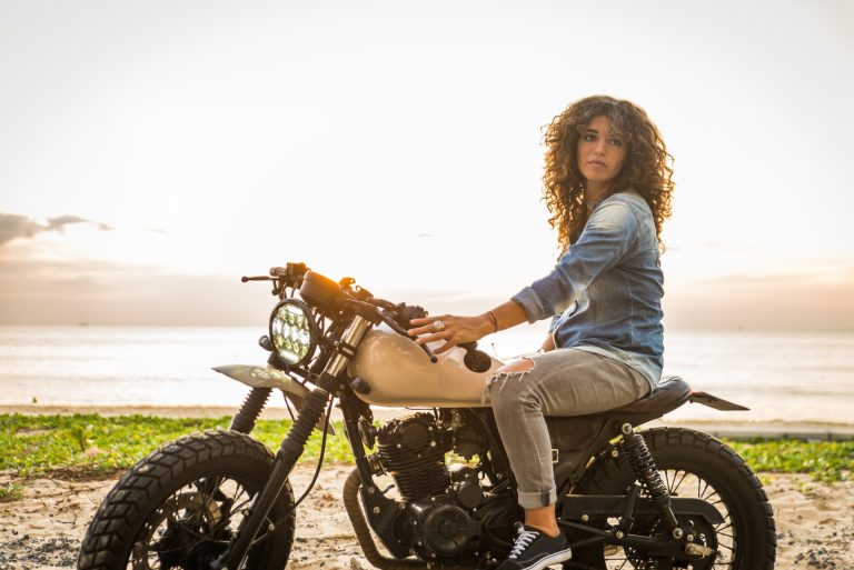 A woman with curly hair sits on a motorcycle parked on a sandy beach at sunset, looking toward the camera.