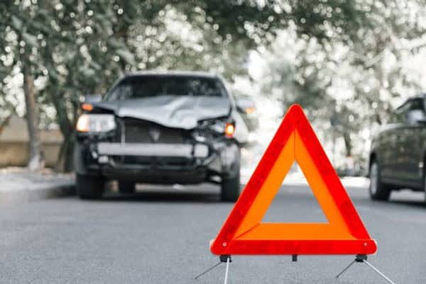 A warning triangle is placed on the road in front of a car with a damaged front end, indicating a recent accident.