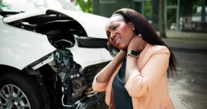 A woman stands in front of a damaged car, holding her neck and appearing to be in pain after an accident.