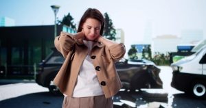 Woman holding her neck in discomfort stands in front of two vehicles involved in a car accident on a city street.