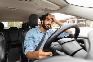 A man in a denim shirt sits in the driver’s seat of a car with one hand on the steering wheel and the other on his forehead, appearing stressed or frustrated.