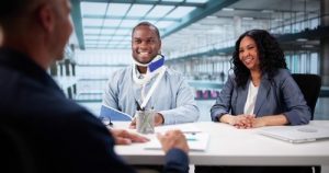 A man with an arm sling and neck brace sits at a desk with a woman, smiling, across from another person in a modern office setting.