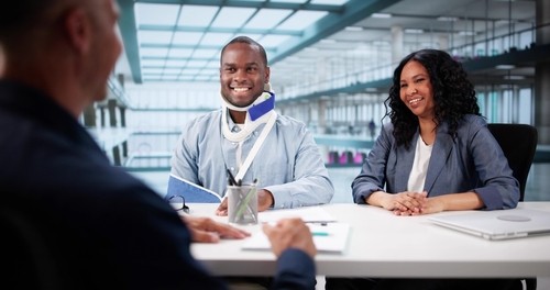 A man with an arm sling and neck brace sits at a desk with a woman, smiling, across from another person in a modern office setting.