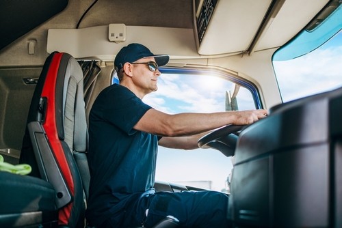 A man wearing sunglasses and a cap drives a truck, viewed from inside the cab with sunlight streaming through the window.